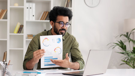 A young Indian male businessman is sitting at the table, talking on a video call on a laptop with colleagues, showing graphs, discussing work points.の写真素材