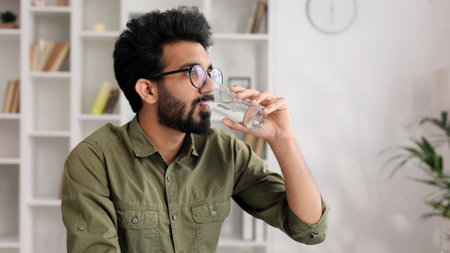 A young Indian boy sits at the table in his living room and starts a new day with a good habit, holds a glass of clean water and drinks it, closeup view.の写真素材