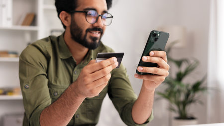 Smiling pretty young Indian brunette boy holding mobile phone and credit card sitting at the table. Happy man makes online shopping using mobile payment. E-banking app service.の写真素材