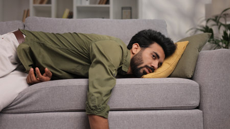 Tired Indian boy slumps on couch feeling listless. Feeling depressed, lack of motivation, sadness, boredom. Exhausted, he collapses on the couch from stress. Burn out.の写真素材