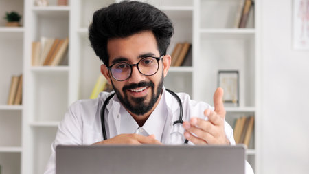 Smart friendly male Indian clinician in white coat using laptop for remote patient consultation in hospital office. Telemedicine. Online appointment with a doctor.の写真素材