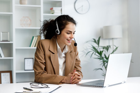 Young curly brunette woman wearing video call headset on laptop in home light office. Video conference, remote meeting. Programmer, remote work, work from home.の写真素材