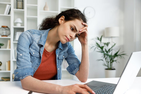 Young curly brunette, sitting stressed at the table and working on a laptop in a bright home office, apathy and depression from overwork at work. Burnout from the modern world. Programmer, studentの写真素材