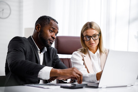 Two people analyze documents on a laptop while sitting at a table in the office. Business partners at work in the office are discussing projects. Multi-racial collaboration.の写真素材