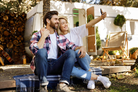 A lovely couple are sitting near the travel trailer, taking a selfie, laughing having fun, enjoying their vacation. Portrait of in love enjoying a sunny day outside the city, drinking wine.の写真素材