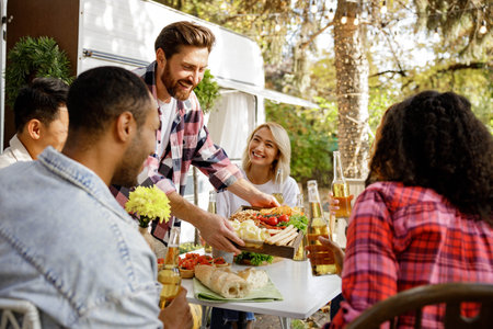 A cheerful multinational company is having fun on a picnic. Multisex friends sitting at the table outdoors celebrating eating and drinking drinks.の写真素材