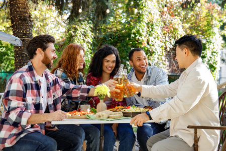A cheerful multinational company is having fun on a picnic. Multisex friends sitting at the table outdoors celebrating eating and drinking drinks.の写真素材