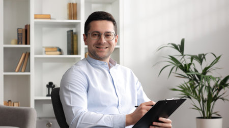 Happy professional psychologist man sitting in chair writing on clipboard, looking and smiling at camera while working in modern office. Psychotherapeutic services, mental health professional concept.の写真素材