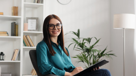 Happy professional psychologist woman sitting in chair writing on clipboard, looking and smiling at camera, working in modern office. Psychotherapeutic services, mental health professional concept.の写真素材