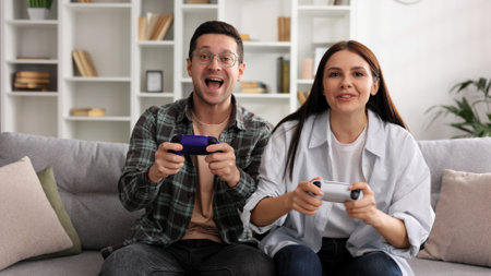 A boy and a girl are having fun playing video games with joysticks in the living room while sitting on the sofa. A couple in love on a date, enjoying the game, joking and having fun.の写真素材