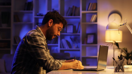 A young Arab student works on his laptop in the evening in the living room. A man with a beard works in IT from home, taps on the keyboard, writes in a notebook.の写真素材