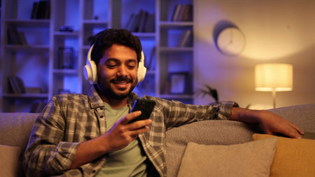 Happy young man with white headphones chilling and listening his favorite song. Arabian brunette man seat on sofa and relax after work, uses smartphone for play music, clicking on the screen.の写真素材