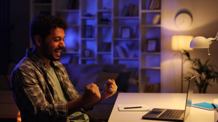 A young man with a beard is happy about winning the lottery in his living room at the table. Happy guy checking email and happy with good news.の写真素材