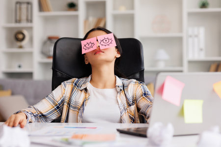 The concept of a lot of work. Tired Indian student girl sleeping with stickers on her eyes while sitting at the table. Distracted woman in home office.の写真素材