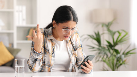 Worried young Indian girl sitting at the table looking at her phone and does not understand why it is not working. The student adjusts her glasses because she does not believe the bad news she saw.の写真素材