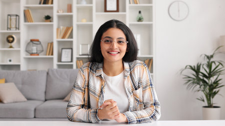 Beautiful Indian girl sitting at the table smiling and looking at the camera. Portrait of a young adult female student in glasses, closeup view.の写真素材
