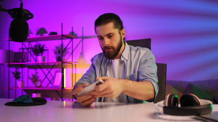 Gamer in neon room. A bearded guy sits at a table and plays a video game on his smartphone. Mobile video games. Rest and relaxation at home.の写真素材