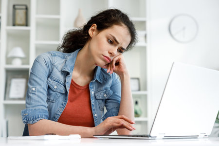 Young curly brunette, sitting stressed at the table and working on a laptop in a bright home office, apathy and depression from overwork at work. Burnout from the modern world. Programmer, studentの写真素材