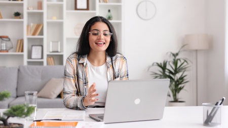 A young Indian girl works at a laptop and looks at the monitor. Beautiful woman talking on video call smiling. Studying in quarantine, listening to a podcast.の写真素材