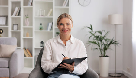 Happy professional psychologist woman sitting in chair writing on clipboard, looking and smiling at camera, working in modern office. Psychotherapeutic services, mental health professional concept.の写真素材