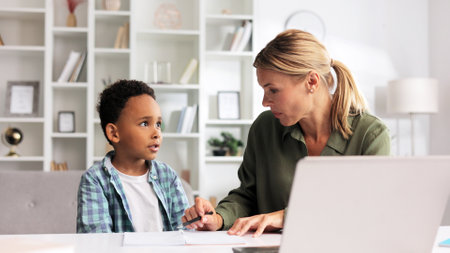 Blonde tutor teaching little African American preschooler to write at home. Help with early childhood education in an online school. Happy boy is learning to write.の写真素材
