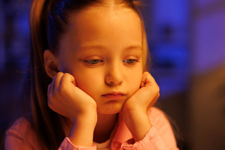 A little girl sits sadly at the table in her living room in the evening. Tired of studying, negative emotions, quarrel with parents, bad grades at school.の写真素材