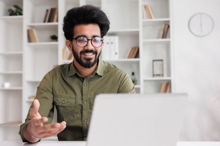 A young Indian male businessman is sitting at the table, talking on a video call on a laptop with colleagues, showing graphs, discussing work points.の写真素材
