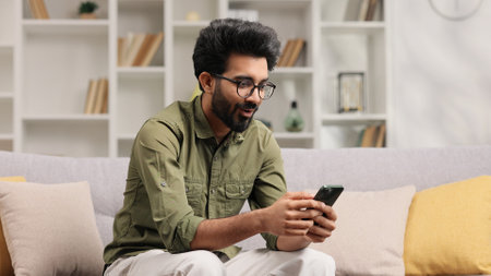 Satisfied Indian man in black glasses checking his email and happy with good news. A young man winner celebrating the victory sitting on the couch, raises his hands uphill.の写真素材