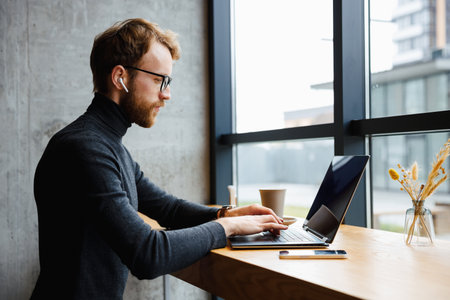 A young red-haired guy, a programmer or entrepreneur in glasses, works in a stylish cafe behind a laptop. Freelancer works remotely. Online communication. Small business.の写真素材