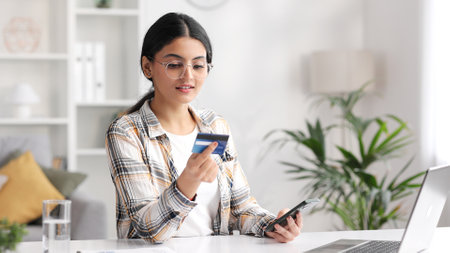 Smiling pretty young Indian brunette girl holding mobile phone and credit card sitting at the table. Happy woman makes online shopping using mobile payment. E-banking app service.の写真素材