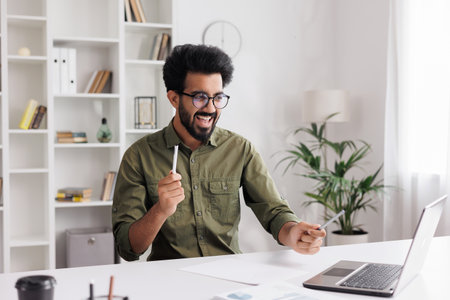 Satisfied Indian guy in black glasses checking his email and happy with good news. A young man winner dancing at the table celebrating the victory, waving pens in hands.の写真素材
