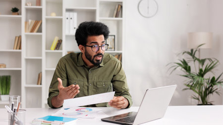 A young Indian man businessman sits at the table, argues on video call on laptop with colleagues, shows graphs, discusses work points, is angry disappointed that he is not listened to or understood.の写真素材