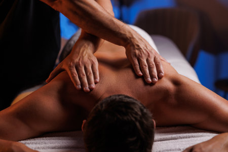 Physiotherapist massaging male patient with damaged shoulder muscle, treating sports injuries. Relaxing professional shoulder massage in cozy atmosphere, reboot on the weekend, end of difficult week.の写真素材