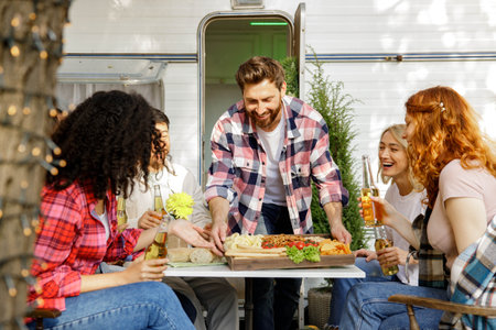 A cheerful multinational company is having fun on a picnic. Multisex friends sitting at the table outdoors celebrating eating and drinking drinks.の写真素材