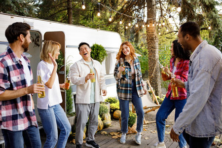 A cheerful group of friends rest in nature near a tourist trailer. Mixed-gender and multinational company of friends toasting, dancing, drinking drinks, have fun.の写真素材