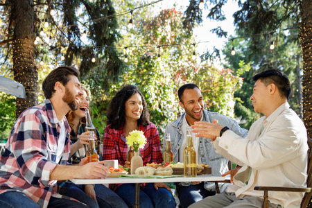 A cheerful multinational company is having fun on a picnic. Multisex friends sitting at the table outdoors celebrating eating and drinking drinks.の写真素材