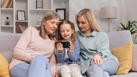 Grayhaired woman grandmother, little granddaughter and blonde mother sit in the living room watching...