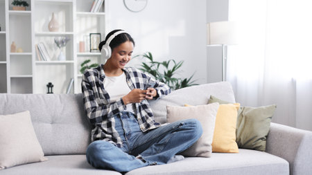Happy young girl with white headphones chilling and listening her favorite song. African American brunette woman sitting on sofa relaxing after work, using smartphone to play music tapping on screen.の写真素材