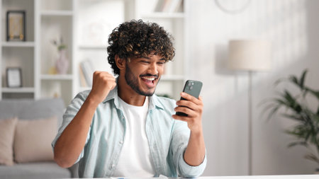 Satisfied Indian man checking his email and happy with good news. A young man winner celebrating the victory sitting at the table, raises his hands uphill.の写真素材