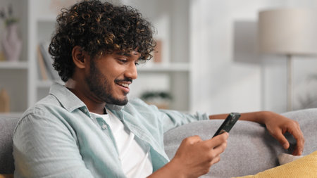 Happy young man with beard chilling and listening his favorite song. Indian brunette man seat on sofa and relax after work, uses smartphone for texting to friends, clicking on the screen.の写真素材