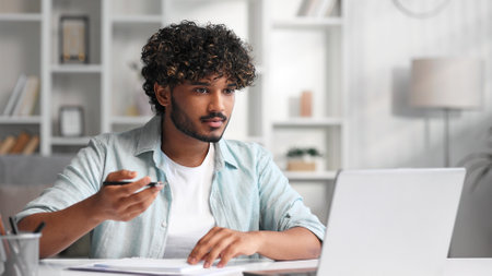 Concentrated Indian male businessman with a beard is studying at a laptop in his living room in the evening. A young guy listens carefully to a video conference, writes down information in a notebook.の写真素材
