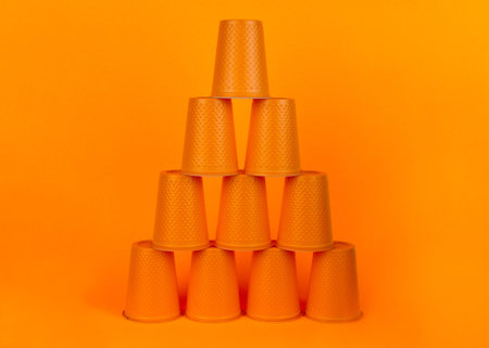 Pyramid of orange plastic disposable cups on an orange background. Waste concept. Minimal space.