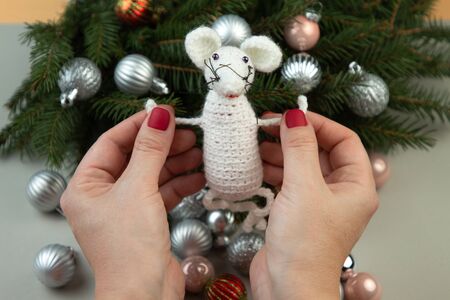 Handmade crochet toy. Amigurumi white rat toy in hands on the background of the Christmas tree. Christmas rat is a symbol of the new year 2020. Chinese New Year.の写真素材