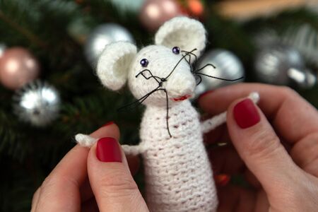 Handmade crochet toy. Amigurumi white rat toy in hands on the background of the Christmas tree. Christmas rat is a symbol of the new year 2020. Chinese New Year.の写真素材