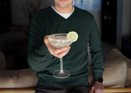 A young man in a green jumper holds a glass with a Margarita cocktail. Gatsby Party. National Margarita Day. Selective focus on a glass with salt and lime. No face.の写真素材
