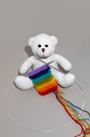 White teddy bear sits on a gray background. Updating old favorite toys. Knitting a rainbow scarf for a teddy bear.の写真素材