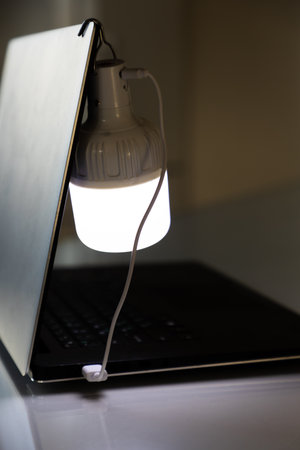 Portable rechargeable LED emergency light with built-in battery is charged from a laptop using a USB cable. Light source during blackout. vertical shot.の写真素材