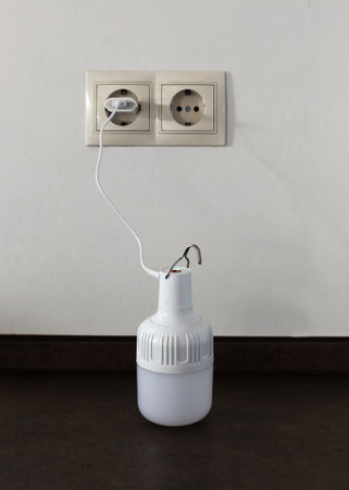 The portable rechargeable LED emergency light with built-in battery is charged from a 220V outlet using a USB cable and adapter. Light source during blackout. vertical shot.の写真素材