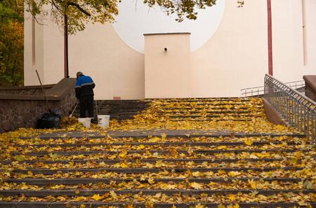 a janitor in overalls removes leaves in garbage bags with a broom and dustpan in the park on the stepsの写真素材