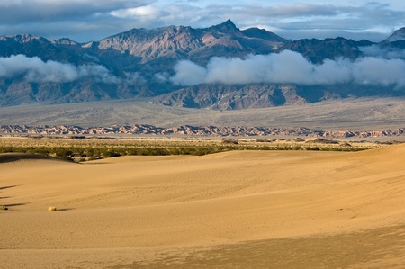 The sun rises on Mesquite Flat Sand Dunes under layers of sunlit mountains and clouds in Death Valley National Park, California.の写真素材
